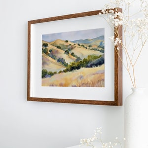 May include: A framed watercolor painting of a landscape with rolling hills, green trees, and a light blue sky. The frame is brown wood.