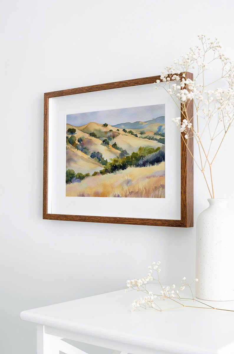 May include: A framed watercolor painting of a landscape with rolling hills, green trees, and a light blue sky. The frame is brown wood.