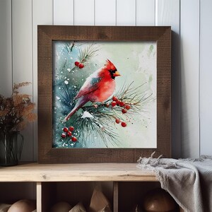 Op de afbeelding: Waterverfschilderij van een rode kardinaal op een dennentak met rode bessen, tegen een besneeuwde, lichtblauwe achtergrond. Het kunstwerk is ingelijst in een donkerbruine houten lijst. De schildering wordt tentoongesteld op een houten plank.