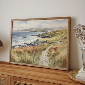 May include: A watercolour painting depicting a coastal scene with a white house on a cliff overlooking a rocky beach and a winding path leading to it.