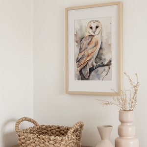 May include: A framed watercolor painting of a barn owl perched on a branch. The owl is brown and white with yellow eyes. The painting is on a white wall with a wicker basket and two ceramic vases on a wooden table.