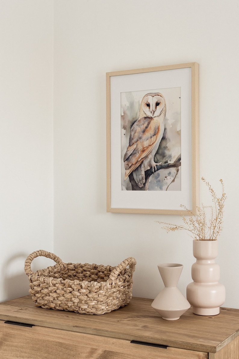 May include: A framed watercolor painting of a barn owl perched on a branch. The owl is brown and white with yellow eyes. The painting is on a white wall with a wicker basket and two ceramic vases on a wooden table.