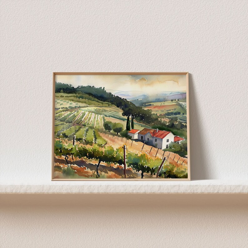May include: Watercolor painting of a vineyard landscape with rolling hills, rows of grapevines, and a farmhouse with a red roof. The artwork is framed in a light brown wooden frame and displayed on a white shelf.