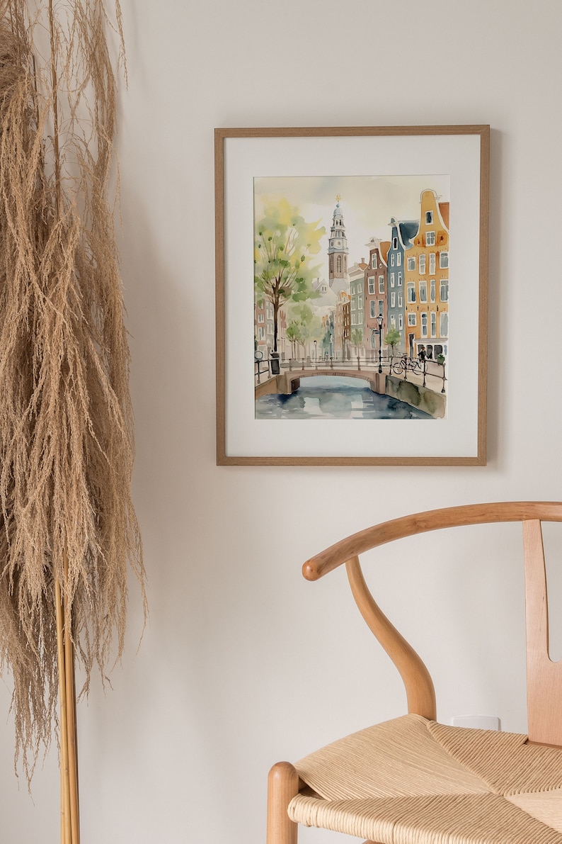 May include: A framed watercolor painting of a canal scene in Amsterdam, Netherlands. The painting features a bridge over a canal with buildings lining the sides. The buildings are painted in a variety of colors, including red, orange, yellow, and blue. There is a tree on the left side of the painting and a bicycle on the bridge.