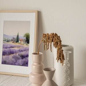 May include: A framed watercolor painting of a lavender field with a small house in the distance. The painting is on a white table with three vases, one pink, one white, and one clear glass vase with a dried flower arrangement.