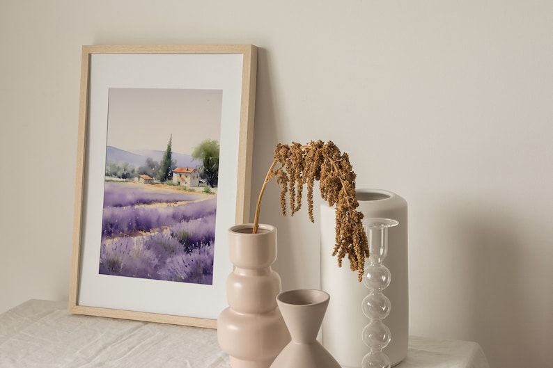 May include: A framed watercolor painting of a lavender field with a small house in the distance. The painting is on a white table with three vases, one pink, one white, and one clear glass vase with a dried flower arrangement.