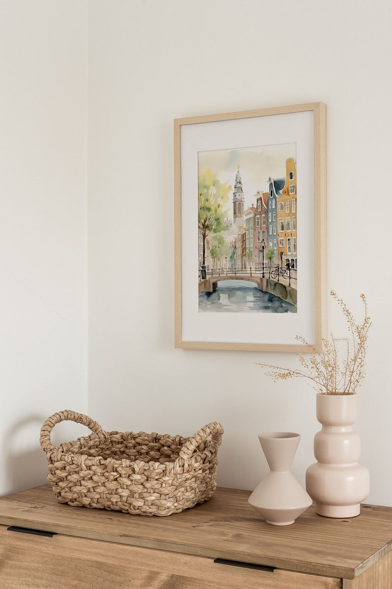 May include: A framed watercolor painting of a canal scene in Amsterdam, Netherlands. The painting features a bridge, buildings, and a bicycle. The painting is hanging on a white wall above a wooden dresser with a woven basket and two ceramic vases.