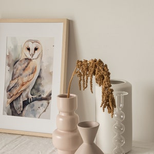 May include: A framed watercolor painting of a barn owl perched on a branch. The owl has brown and white feathers and yellow eyes. The painting is displayed on a white table with a vase of dried flowers and two other vases.