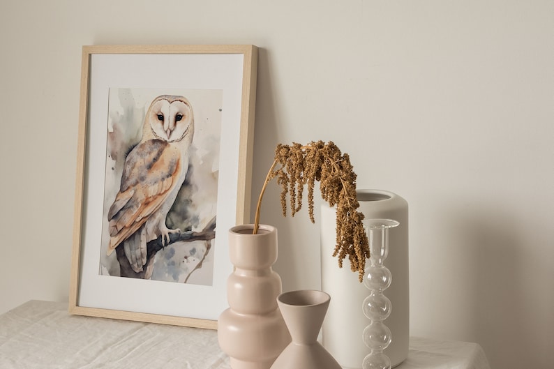 May include: A framed watercolor painting of a barn owl perched on a branch. The owl has brown and white feathers and yellow eyes. The painting is displayed on a white table with a vase of dried flowers and two other vases.