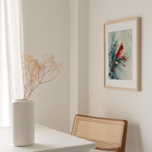 Op de afbeelding: Een ingelijste aquarelprint van een rode kardinaal die op een tak met rode bessen zit. Het kunstwerk wordt tentoongesteld aan een witte muur, naast een witte tafel met een vaas en een houten stoel met een geweven rug.