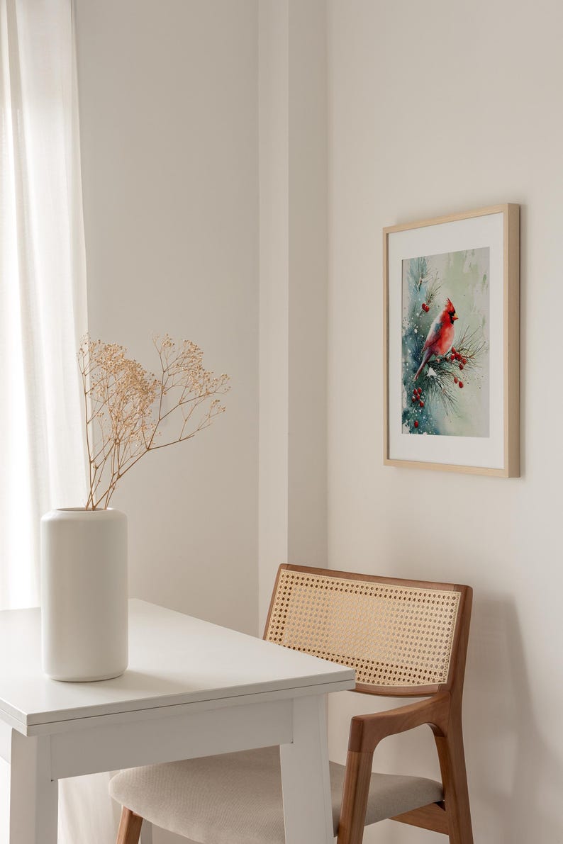 Op de afbeelding: Een ingelijste aquarelprint van een rode kardinaal die op een tak met rode bessen zit. Het kunstwerk wordt tentoongesteld aan een witte muur, naast een witte tafel met een vaas en een houten stoel met een geweven rug.