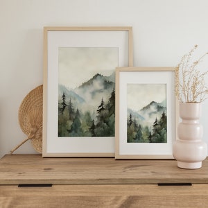 Pu&ograve; includere: Due dipinti ad acquerello incorniciati di un paesaggio montano nebbioso con alberi verdi. I dipinti sono in cornici di legno naturale e sono esposti su una cassettiera in legno.