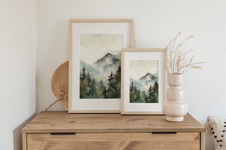 Pu&ograve; includere: Due dipinti ad acquerello incorniciati di un paesaggio montano nebbioso con alberi verdi. I dipinti sono in cornici di legno naturale e sono esposti su una cassettiera in legno.