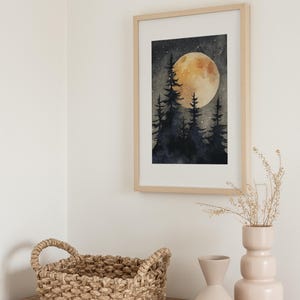 May include: A framed watercolor print depicts a nighttime forest scene with silhouetted trees and a large, glowing moon. The artwork is in a light wood frame, displayed above a wooden surface with a woven basket and two decorative vases.