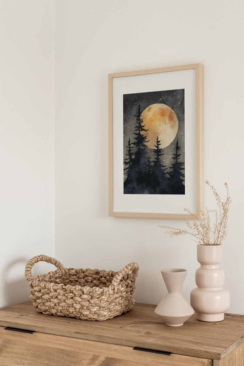 May include: A framed watercolor print depicts a nighttime forest scene with silhouetted trees and a large, glowing moon. The artwork is in a light wood frame, displayed above a wooden surface with a woven basket and two decorative vases.
