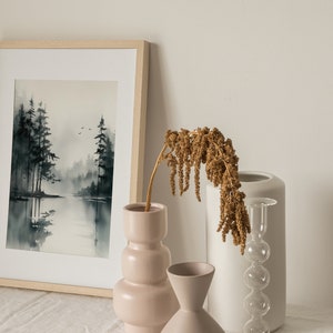 May include: A framed watercolor painting of a misty forest scene with a lake in the foreground. The painting is displayed on a table with three decorative vases, one pink, one white, and one clear glass with a bulbous shape. A dried flower arrangement is in the pink vase.