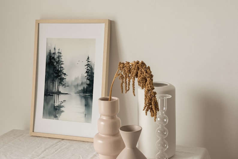 May include: A framed watercolor painting of a misty forest scene with a lake in the foreground. The painting is displayed on a table with three decorative vases, one pink, one white, and one clear glass with a bulbous shape. A dried flower arrangement is in the pink vase.