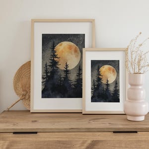 May include: Two framed watercolor prints of a night forest scene with a large, glowing moon. The artwork features dark blue trees and a starry sky. The frames are light wood, and the prints are displayed on a wooden dresser.