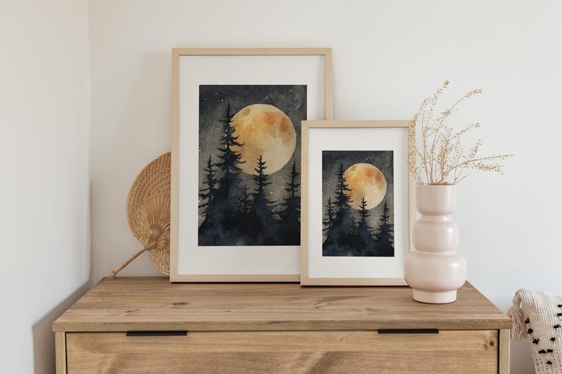 May include: Two framed watercolor prints of a night forest scene with a large, glowing moon. The artwork features dark blue trees and a starry sky. The frames are light wood, and the prints are displayed on a wooden dresser.