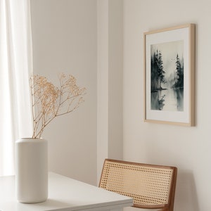 May include: A white table with a white vase filled with dried flowers sits next to a wooden chair with a woven cane seat. A framed black and white watercolor painting of a forest scene hangs on the wall behind the chair.