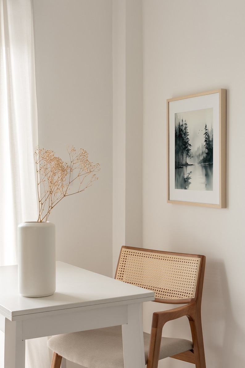 May include: A white table with a white vase filled with dried flowers sits next to a wooden chair with a woven cane seat. A framed black and white watercolor painting of a forest scene hangs on the wall behind the chair.