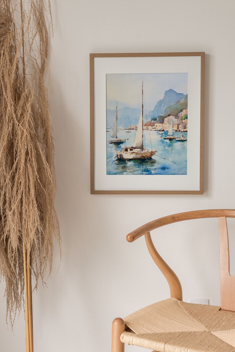 May include: A framed watercolour painting of a sailboat with white sails on a body of water with other sailboats in the distance. The painting is set against a backdrop of a mountain range and a town.