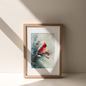 Op de afbeelding: Waterverfschilderij van een rode kardinaal die op een tak zit met rode bessen en groene dennennaalden. Het kunstwerk is ingelijst in een lichtbruine houten lijst met een witte passe-partout. Het thema is natuur en winter.
