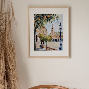 May include: A watercolour painting of a Spanish plaza with a clock tower, orange trees, and a street lamp. The painting is framed in a light wood frame.