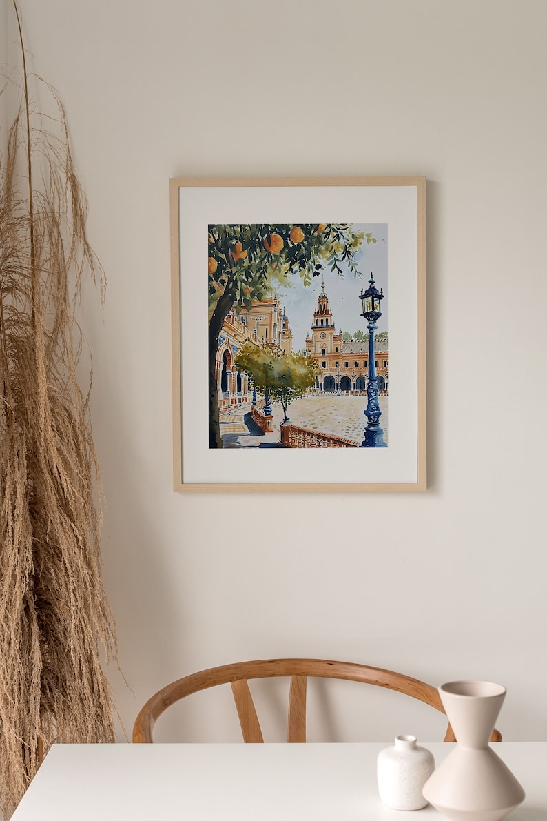 May include: A watercolour painting of a Spanish plaza with a clock tower, orange trees, and a street lamp. The painting is framed in a light wood frame.