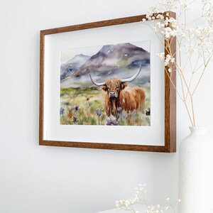 May include: A watercolor painting of a Highland cow standing in a field of wildflowers with a mountain range in the background. The cow is brown with long horns and a fluffy mane. The painting is framed in a brown wood frame.