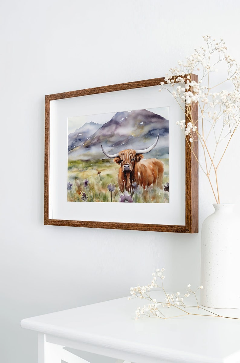 May include: A watercolor painting of a Highland cow standing in a field of wildflowers with a mountain range in the background. The cow is brown with long horns and a fluffy mane. The painting is framed in a brown wood frame.