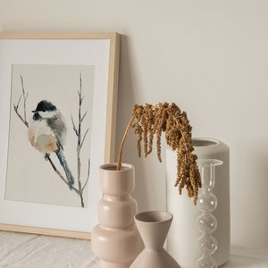 Peut inclure: Une peinture &agrave; l'aquarelle encadr&eacute;e d'un petit oiseau perch&eacute; sur une branche, expos&eacute;e sur une table avec trois vases. Deux des vases sont en c&eacute;ramique, l'un est rose et l'autre est blanc. Le troisi&egrave;me vase est en verre transparent avec une forme bulbeuse. Un arrangement de fleurs s&eacute;ch&eacute;es est dans le vase rose.