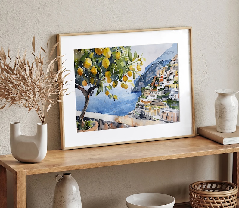 May include: A framed watercolor painting of a lemon tree with yellow lemons and green leaves in front of a colorful cityscape on a cliff overlooking a blue body of water.