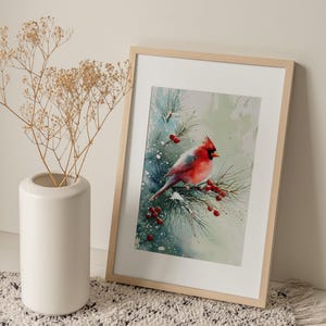 Op de afbeelding: Een ingelijste aquarelprint van een rode kardinaal die op een met sneeuw bedekte dennentak met rode bessen zit. Het kunstwerk staat in een licht houten lijst, leunend tegen een muur. Links staat een witte vaas met gedroogde planten.