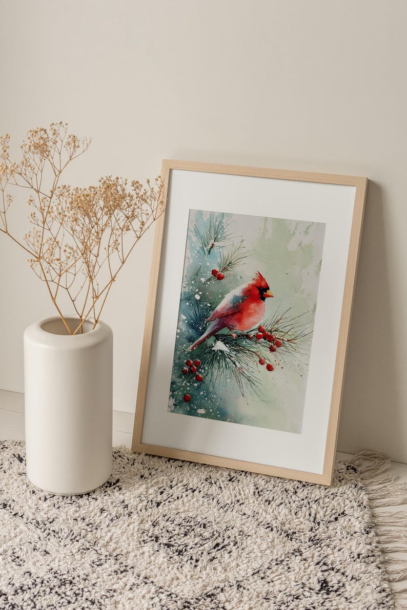 Op de afbeelding: Een ingelijste aquarelprint van een rode kardinaal die op een met sneeuw bedekte dennentak met rode bessen zit. Het kunstwerk staat in een licht houten lijst, leunend tegen een muur. Links staat een witte vaas met gedroogde planten.