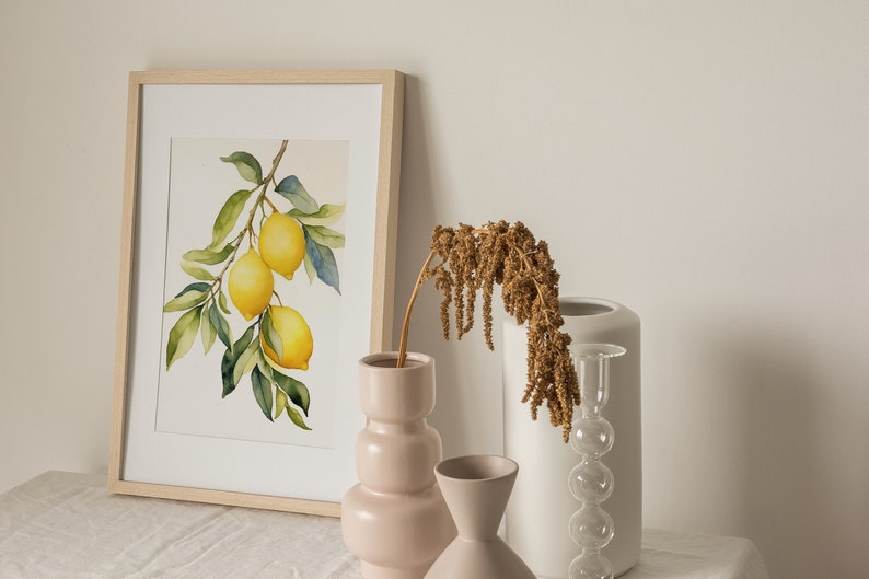 Op de afbeelding: Een ingelijst aquarel schilderij van een citroen tak met drie citroenen die eraan hangen. Het schilderij staat op een witte tafel met drie vazen, een roze, een witte en een heldere glazen vaas met een bruine gedroogde bloemen arrangement.