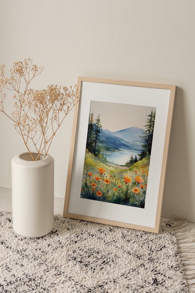 Puede incluir: Una pintura de acuarela enmarcada de una escena de lago de monta&ntilde;a con amapolas naranjas en primer plano. El lago est&aacute; rodeado de &aacute;rboles y monta&ntilde;as. La pintura est&aacute; en un marco de madera claro.