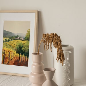 Puede incluir: Una pintura de acuarela enmarcada de un paisaje de vi&ntilde;edo con una granja en la distancia. La pintura se exhibe en una mesa blanca con tres jarrones, uno con flores secas, uno blanco y uno rosa.