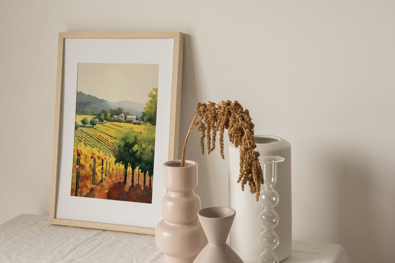 Puede incluir: Una pintura de acuarela enmarcada de un paisaje de vi&ntilde;edo con una granja en la distancia. La pintura se exhibe en una mesa blanca con tres jarrones, uno con flores secas, uno blanco y uno rosa.