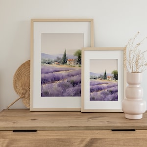 May include: Two framed watercolor paintings of a lavender field with a small house in the distance. The paintings are hanging on a light wood wall above a wooden dresser.