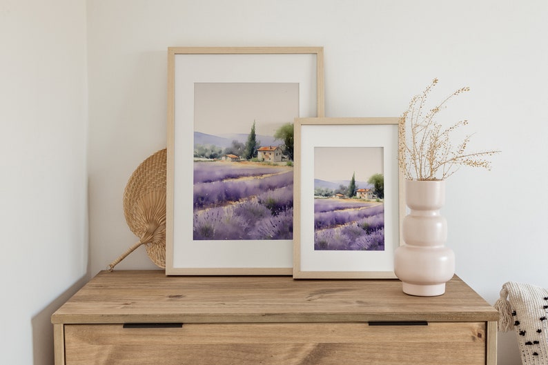 May include: Two framed watercolor paintings of a lavender field with a small house in the distance. The paintings are hanging on a light wood wall above a wooden dresser.
