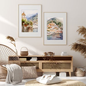 May include: Two framed watercolor paintings of a coastal town with buildings, a church, and a boat in the water. The paintings are hanging on a white wall above a wooden cabinet with a wicker chair and a throw blanket.