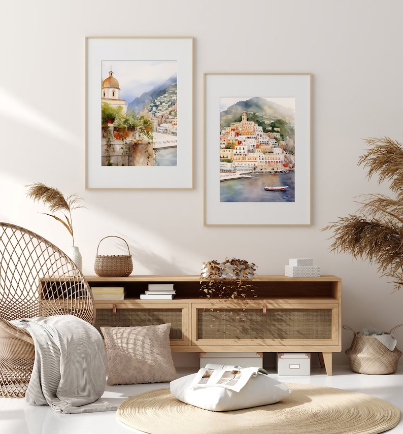 May include: Two framed watercolor paintings of a coastal town with buildings, a church, and a boat in the water. The paintings are hanging on a white wall above a wooden cabinet with a wicker chair and a throw blanket.