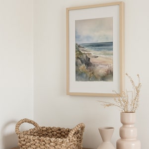 May include: A framed watercolor painting of a beach scene with a light blue sky, a sandy beach, and a rocky cliff. The painting is hanging on a white wall above a wooden dresser with a woven basket and two ceramic vases.
