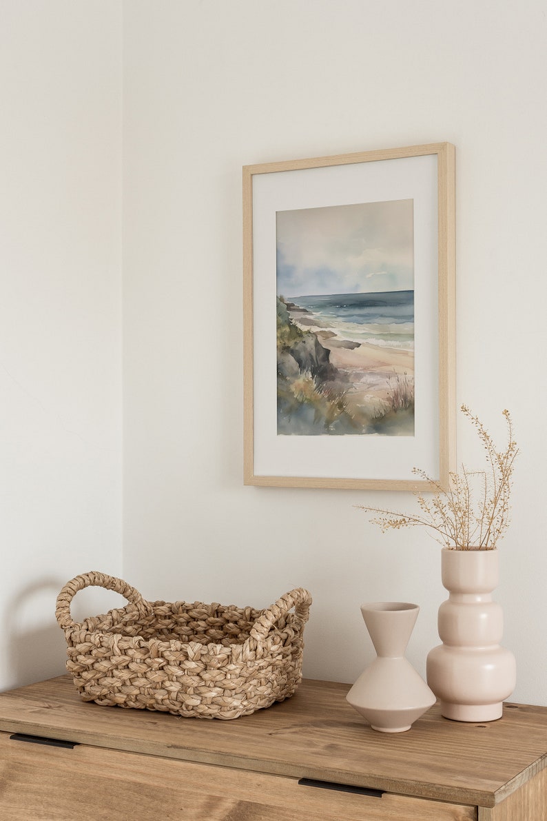May include: A framed watercolor painting of a beach scene with a light blue sky, a sandy beach, and a rocky cliff. The painting is hanging on a white wall above a wooden dresser with a woven basket and two ceramic vases.