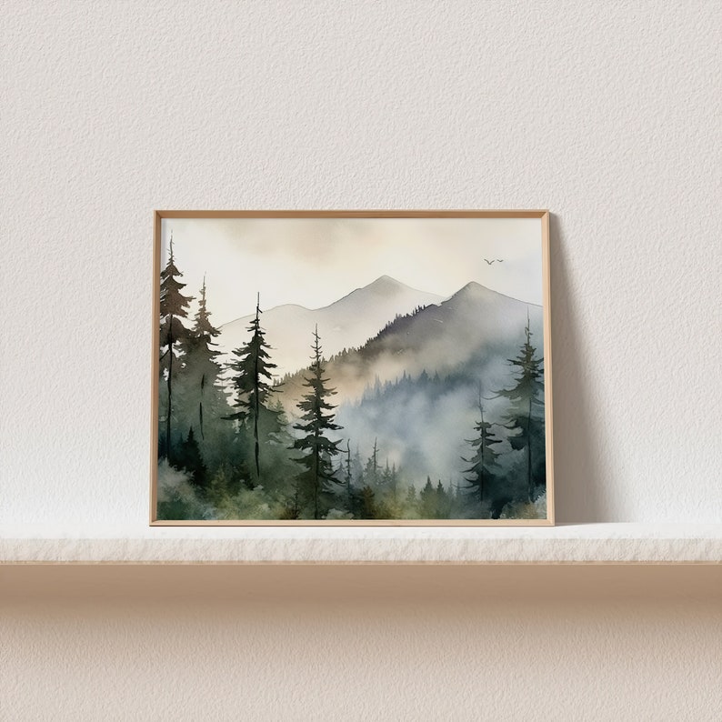 May include: A watercolor painting of a misty mountain landscape with a forest of evergreen trees in the foreground. The sky is a soft, muted yellow, and the mountains are a hazy blue-gray. The painting is framed in a simple, light wood frame.