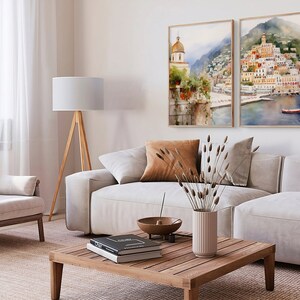 May include: Two watercolor paintings of a coastal town with buildings, a church, and a boat in the water. The paintings are framed in wood and hung on a white wall above a light gray couch and a wooden coffee table.