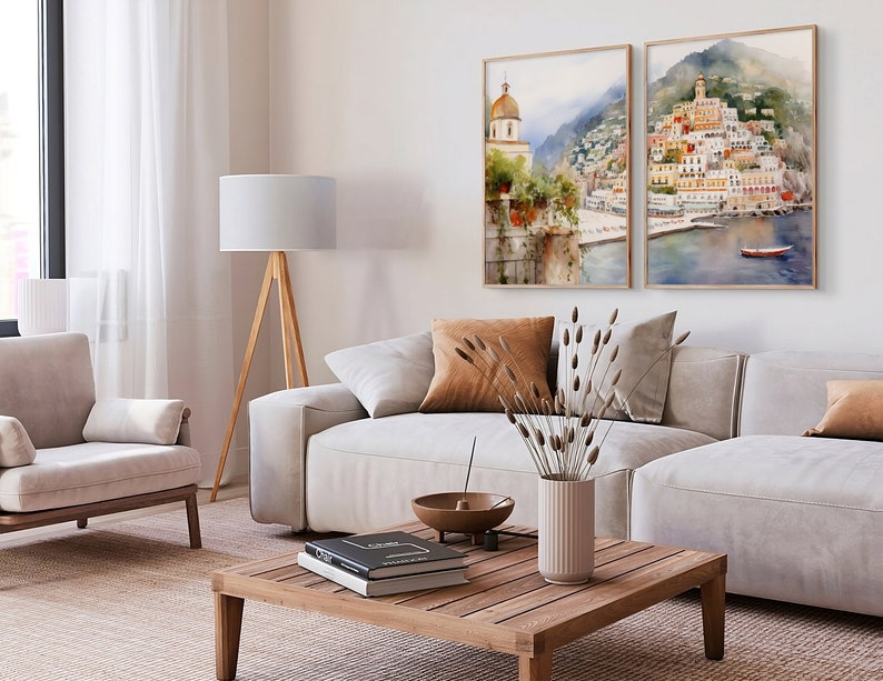 May include: Two watercolor paintings of a coastal town with buildings, a church, and a boat in the water. The paintings are framed in wood and hung on a white wall above a light gray couch and a wooden coffee table.
