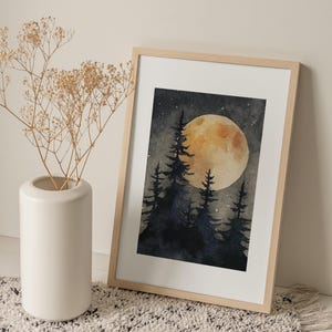 May include: Watercolor painting of a forest scene with a large, yellow moon and silhouetted trees. The artwork is framed in a light wood frame and leans against a wall. A white vase with dried flowers sits to the left.
