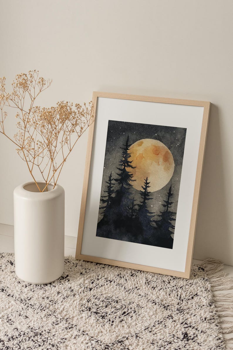 May include: Watercolor painting of a forest scene with a large, yellow moon and silhouetted trees. The artwork is framed in a light wood frame and leans against a wall. A white vase with dried flowers sits to the left.